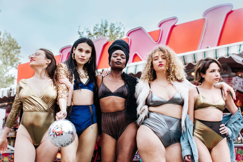 Cinq femmes aux corps variés, en lingerie métallisée, posent bras dessus bras dessous devant un manège de fête foraine